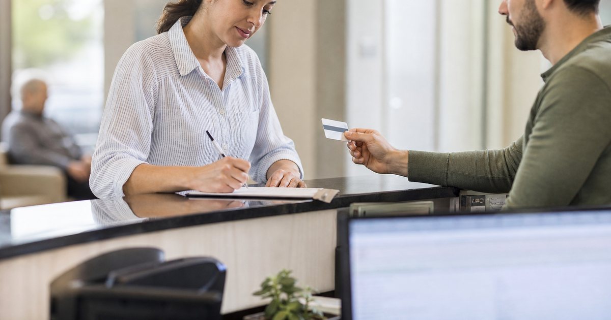 A front desk assistant at a family clinic updating a patient's details on a paper form while the patient holds up an insurance card, daytime