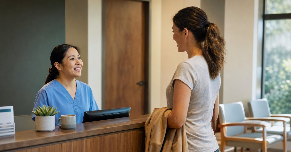 A sunlit clinic reception counter in warm morning light, a receptionist in light blue scrubs smiling at a patient who stands with a jacket folded over one arm, wooden desk with a ceramic mug and a small potted plant, blurred empty waiting room chairs behind
